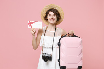 Smiling young tourist girl in summer dress hat with photo camera isolated on pink background. Traveling abroad to travel weekend getaway. Air flight journey concept. Hold gift certificate, suitcase.