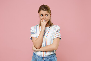 Worried young blonde woman girl in casual striped shirt posing isolated on pastel pink background studio portrait. People emotions lifestyle concept. Mock up copy space. Gnawing nails, looking aside.