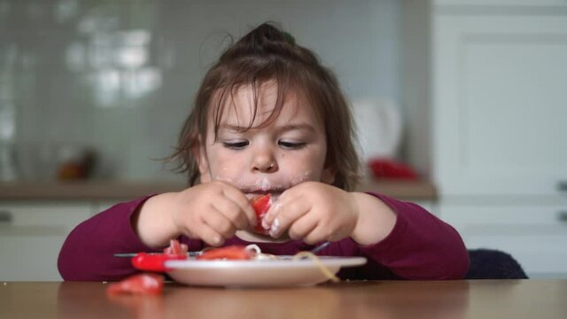toddler girl eats spaghetti with tomatoes and cream at home with hands. messy baby mealtime