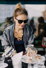 Beautiful girl in black glasses, with gathered hair and ethnic clothes sits in a restaurant on the street and takes a sandwich with a smile.
