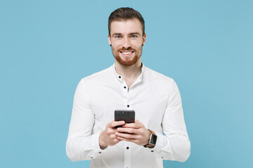 Smiling young bearded man guy 20s in white classic shirt isolated on pastel blue wall background studio portrait. People lifestyle concept. Mock up copy space. Using mobile phone, typing sms message.