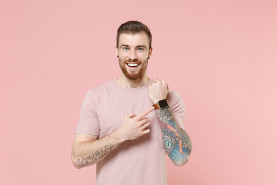 Funny young bearded tattooed man guy in pastel casual t-shirt posing isolated on pink background in studio. People lifestyle concept. Mock up copy space. Pointing index finger on smart watch on hand.