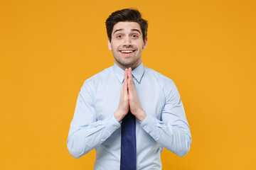 Excited young business man in classic blue shirt tie posing isolated on yellow background studio portrait. Achievement career wealth business concept. Mock up copy space. Hold hands folded in prayer.