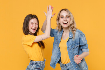 Two cheerful young women girls friends in casual t-shirts denim clothes posing isolated on yellow...