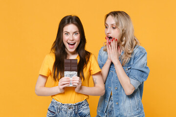 Two excited young women girls friends in casual t-shirts denim clothes isolated on yellow wall background studio. People lifestyle concept. Mock up copy space. Hold chocolate bar, put hands on cheeks.