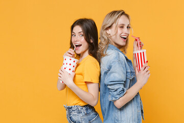 Two excited women girls friends in casual t-shirts denim clothes isolated on yellow background. People lifestyle concept. Mock up copy space. Hold cups of soda or cola, stand back to back, blinking.