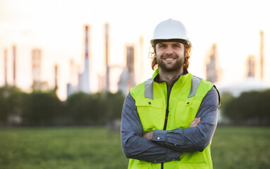 Young engineer standing outdoors by oil refinery, looking at camera.