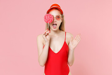 Shocked young blonde woman in red one-piece swimsuit cap eyeglasses isolated on pink background. People summer vacation rest lifestyle concept. Mock up copy space. Covering eye with round lollipop.