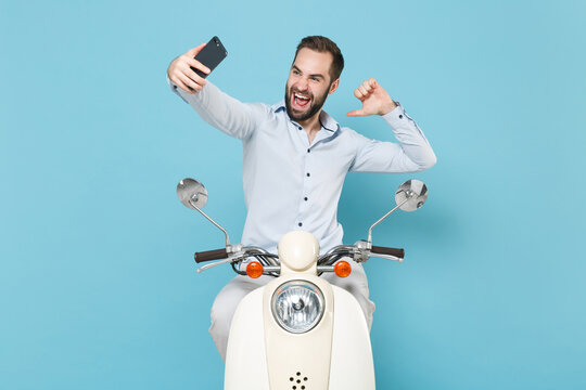 Cheerful young bearded man guy in casual light shirt driving moped isolated on blue background. Driving motorbike transportation concept. Doing selfie shot on mobile phone, pointing thumb on himself.