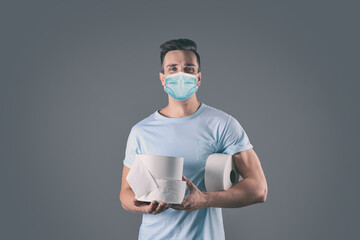 Man in medical mask holding toilet paper rolls on grey background