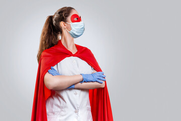 A doctor wearing surgical gloves and a mask, wearing a red superhero Cape, poses with her arms...
