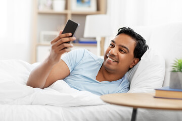 people, bedtime and rest concept - happy smiling indian man with smartphone lying in bed at home