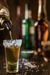 An alcoholic drink at the bar counter is poured into a tequila glass with salt at the edges, in the background are different bottles of alcohol, shallow depth of field, selective focus. The concept of