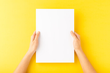 Young woman’s hands showing blank paper sheet on yellow desktop. Top view
