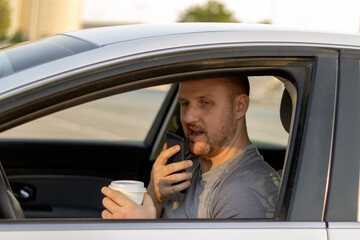 Side view,serious young man in the car while drinking coffee.Close up,sportsman in car using mobile.Handsome bearded male going to work in comfortable car.Man driving,drinking coffee and using phone.