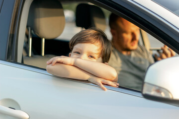Portrait of cute little boy leaning out of car window outside while his father useing...