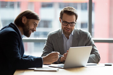 Caucasian teammates two positive businessmen sitting at desk in modern office looking at computer screen using learn new corporate e-business application, modern technology business management concept
