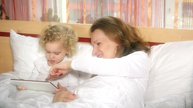 mother and little curly daughter play game video on tablet in bed. Childhood. 4K