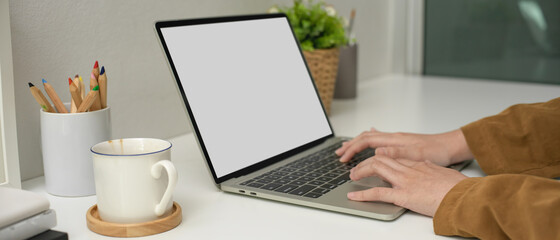 Female employee working with mock-up laptop, cup, coloured pencils and decoration on white desk beside window