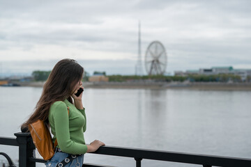 a girl stands on the embankment of the river, looks at the other side and talks on the phone.  on the other side of the river the Chinese city of Heihe, a Ferris wheel.