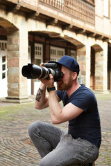 Junger Fotograf mit blauer Kappe hält eine große Kamera DSLR in der Hand mit einem Teleobjektiv, er hat eine Kamera Tattoo auf dem Unterarm