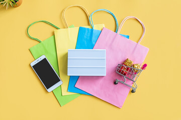 Flat lay of empty lightbox, smartphone, miniature gift boxes in trolley and colorful bags on yellow background. Top view and copy space for text. Online shopping concept.