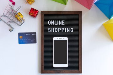 Miniature of gift boxes in trolley, smartphone, word on black frame, credit card and colorful bags on white background. Online shopping, new normal concept. Top view, flat lay and copy space