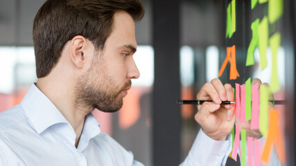 Close up side view focused businessman writing on colorful sticky notes attached on glass wall,...