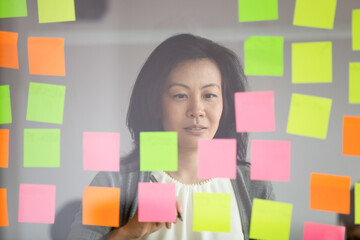Mature asian female coach make presentation for staff attaching sticky notes on glass wall view...