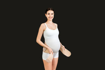 Portrait of pregnant woman dressing maternity belt against pain in the back at black background with copy space. Orthopedic abdominal support belt concept