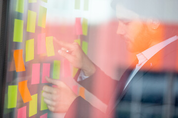 View through window focused successful businessman in suit working on new project attaching on wall colorful sticky notes writing steps of solutions, decision options, producing start up ideas concept