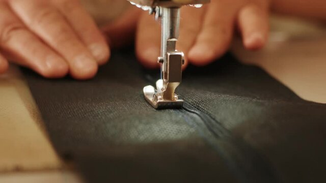 Hand of the seamstress is using white industrial sewing machine sew zipper (slide fastener) of crotch black pants close-up.