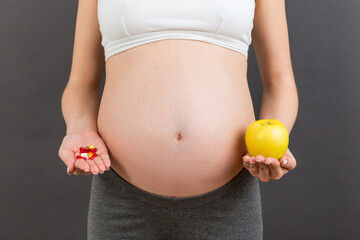 Close up of pregnant woman making a choice between an apple and a pile of pills at colorful background with copy space. Taking vitamins during pregnancy concept