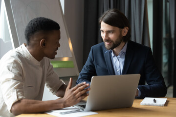 African and Caucasian businessmen talking seated at desk in modern office boardroom, sharing ideas, startup creative thoughts, client and manager friendly communication, sales and mentoring concept