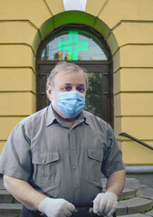 Sad elderly man wearing medical mask and white gloves stands near hospital entrance