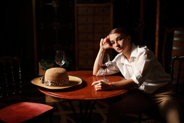 Beautiful theatrical actress woman with retro fashion style on the stage siting near wooden table with glass of red wine and vintage hat. 