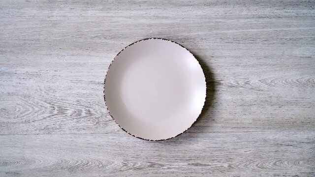 Close-up of an empty white plate on a light wooden background for the design. Lunch dish on top of a tree background. Slow approach of the video to the plate, top view.