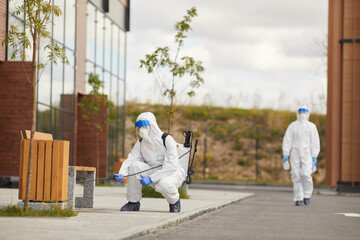 Full length portrait of two workers wearing protective suits spraying chemicals over bench outdoors during disinfection or cleaning, copy space