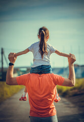 Obraz premium Dad dancing on his shoulders with his daughter in sun. Father travels with baby on his shoulders in rays of sunset. Child with parents walks at sunset. Happy fathers day concept.