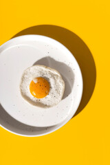 Fried egg on a round plate on a yellow table with hard shadows. The concept of creativity and minimalism in cooking. Top view, flat lay.