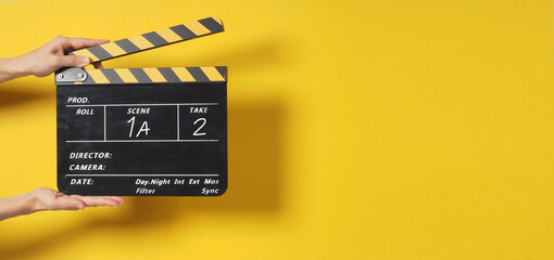 A Hand is holding Black clapperboard or movie slate on yellow background.It has written a number..