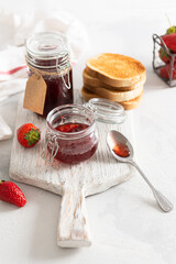 Sweet homemade strawberry jam in glass jars and fresh berries on white wooden board on rustic white background. Side view, close up. Cookbook recipe. Social media content. Summer strawberry confiture