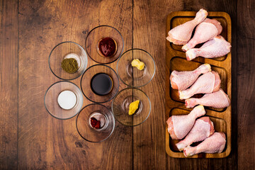A wooden board full of FRESH RAW ORGANIC CHICKEN DRUMSTICK along with lots of glass bowls with different kinds of spices on the table with free space for text/decoration.