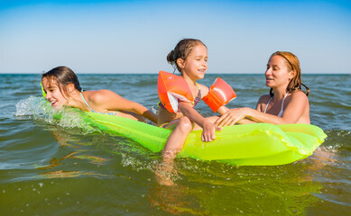Obraz premium Happy mother and two small positive daughters bathe and swim in the sea with an air mattress on a sunny summer day. Vacation concept with kids. Copyspace