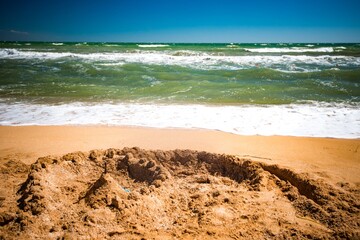 Sand fortress built by children stands on the background of noisy sea waves on a sunny warm summer day. Family travel concept with kids and tourism
