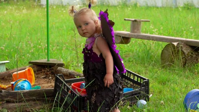 Little cute blond girl with purple butterfly wings plays near the sandbox outdoors