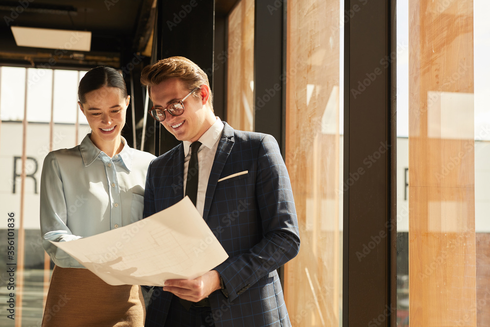 Wall mural Waist up portrait of smiling rental agent showing floor plans to female client while standing in office building interior lit by sunlight, copy space