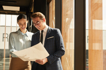Waist up portrait of smiling rental agent showing floor plans to female client while standing in office building interior lit by sunlight, copy space - obrazy, fototapety, plakaty