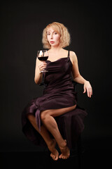 Middle age blonde woman drinking glass of red wine in elegant dress over isolated black background background 
