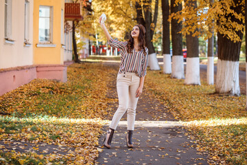 Cheerful and fashionable woman is taking selfie with her instax camera outdoors in autumn. Yellow foliage on and under trees. 